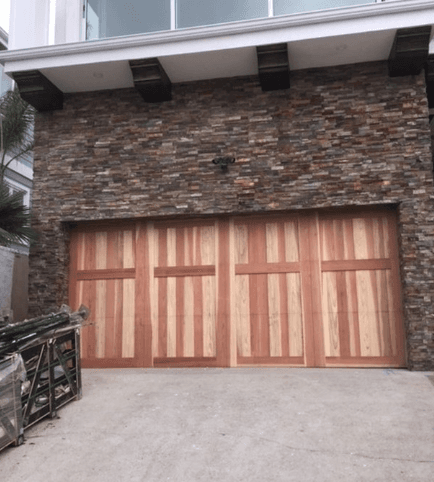 Four-panel natural wood garage door set into a rustic multi-colored stone veneer wall.