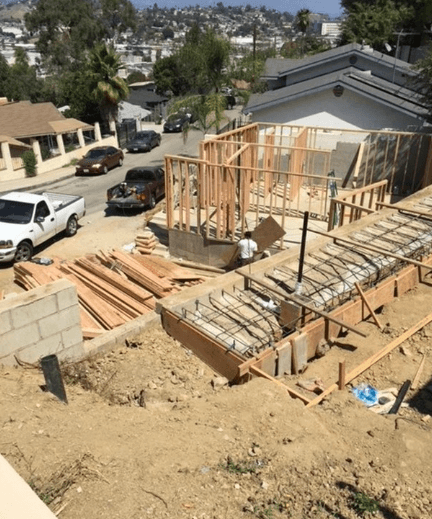Worker carries lumber at a hillside residential construction site with wooden framing and rebar.